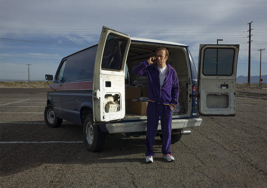 Jimmy McGill (Bob Odenkirk) in a purple track suit, talking on a cell phone while standing in front of a large cargo van with the back open and a number of cardboard boxes inside.